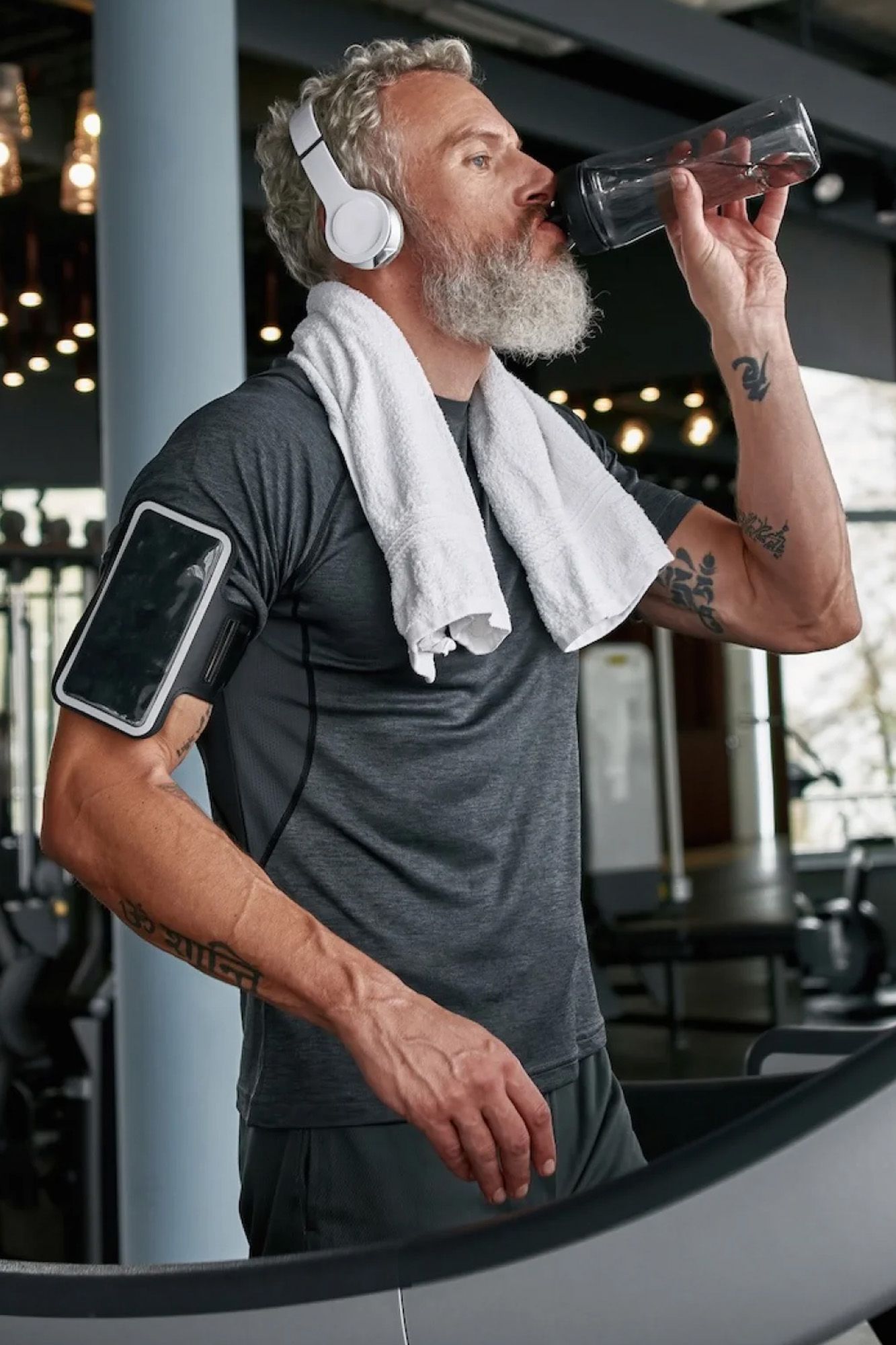 Man at the gym wearing headphones, drinking water, with a towel on his neck and phone strapped to his arm.