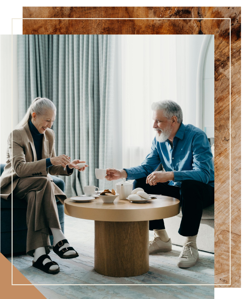 Two elderly men sit at a coffee table, smiling and enjoying tea and snacks together indoors.
