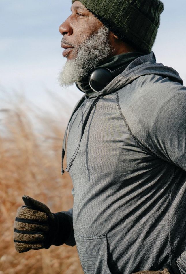 Man in winter clothes jogging outdoors, wearing gloves, hoodie, beanie, and headphones around his neck.