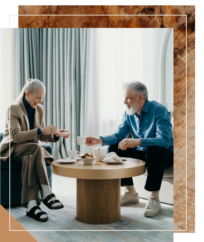 Two elderly men sit at a coffee table, smiling and enjoying tea and snacks together indoors.